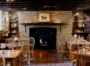 RedFoxInn-Tavern-and-Food_001-19c46a1713b94357af587a369494b45e.webp Cozy dining room at The Red Fox Inn & Tavern with stone fireplace and wooden furniture.